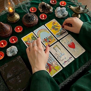 Op de afbeelding: Een verzameling tarotkaarten uitgestald op een donkergroen fluwelen kleed, waaronder de Tien van Kelken, De Dwaas, Drie van Zwaarden en Aas van Zwaarden. De sc&egrave;ne bevat brandende rode theelichtjes, een koperen olielamp, kristallen en een donker dagboek.
