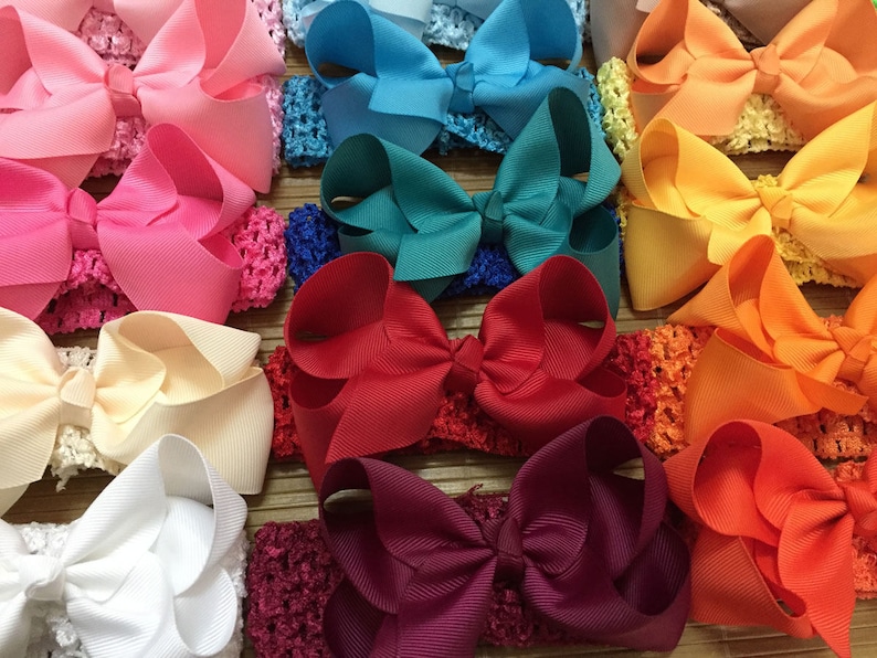 May include: A collection of colourful hair bows with crocheted headbands. The bows are arranged in a rainbow pattern, starting with pink and ending with orange.