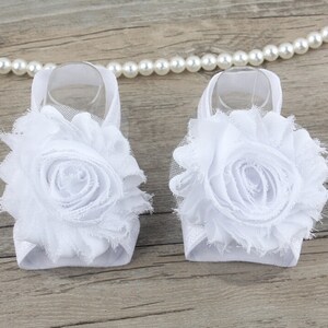 K&ouml;nnte beinhalten: Ein Paar wei&szlig;e Babysandalen mit einer wei&szlig;en Blumenverzierung. Die Sandalen sind aus einem weichen, dehnbaren Material gefertigt und haben ein zartes, feminines Design.