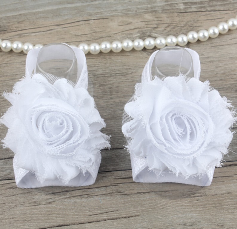 K&ouml;nnte beinhalten: Ein Paar wei&szlig;e Babysandalen mit einer wei&szlig;en Blumenverzierung. Die Sandalen sind aus einem weichen, dehnbaren Material gefertigt und haben ein zartes, feminines Design.