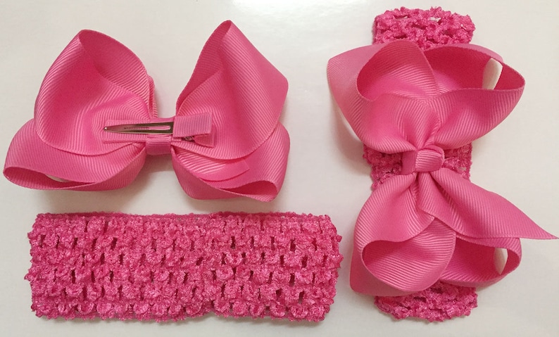 May include: Two pink hair bows, one with a clip and one attached to a pink elastic headband. Both bows are made of a shiny, satin-like material.
