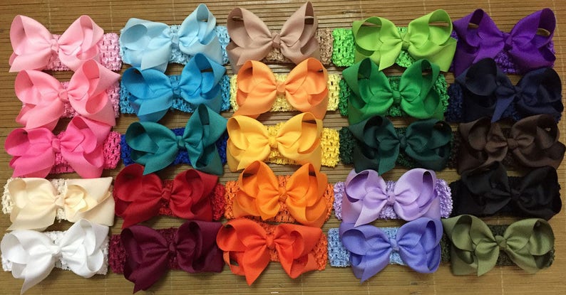 May include: Twenty-four hair bows in a variety of colours, including pink, blue, green, yellow, orange, red, purple, brown, black, and white. Each bow is attached to a crocheted headband.