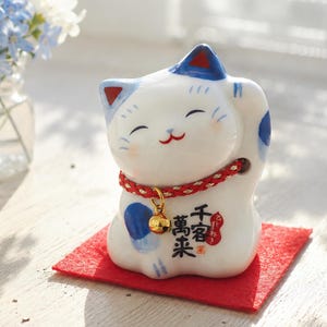 May include: A white ceramic Maneki Neko cat figurine with blue and red details, a red collar, and a gold bell. The cat is on a red felt square. A small glass vase with blue and white flowers is in the background.