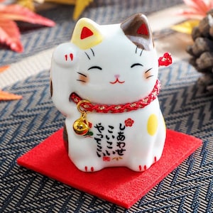 以下が含まれることがあります： 白いセラミック製の招き猫の置物。茶色と黄色のアクセントがあり、赤い首輪に金の鈴が付いています。猫は赤い正方形の上に座っており、胸には日本語のテキストがあります。猫は左手を上げています。