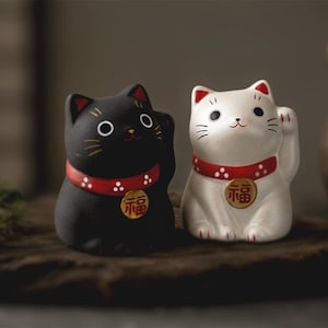 Könnte beinhalten: Zwei Keramik-Katzenfiguren, eine in Mattschwarz und eine in Cremeweiß, jeweils mit einem roten Halsband mit weißen Punkten und einem goldenen runden Anhänger mit dem chinesischen Schriftzeichen für Glück. Beide Katzen haben eine Pfote erhoben.
