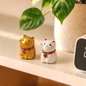 May include: Two small cat figurines, one metallic gold and one white, each featuring a red collar with white dots and a gold-colored circular pendant displaying the black Chinese character for good fortune, known as Fu, on the chest area.