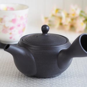 以下が含まれることがあります： 黒いセラミックティーポット。表面はテクスチャ加工され、小さなハンドルが付いています。ピンクの花柄の白いティーカップが、背景の黒いコースターの上にあります。ティーポットには、小さなノブが付いた丸い蓋が付いています。