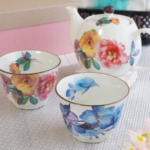 May include: A white ceramic tea set with a teapot and two teacups, decorated with watercolour floral patterns in red, yellow, and blue. The teapot has a lid and a brown knob. The teacups have a brown rim.