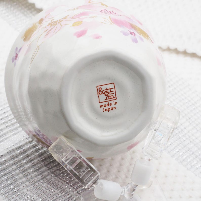 Pu&ograve; includere: Ciotola in ceramica bianca con design floreale rosa e dettagli dorati. La ciotola ha un timbro rosso con la scritta "made in Japan". Clip di plastica trasparente sono attaccate alla ciotola.