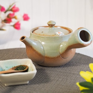 May include: A ceramic teapot with a side handle, spout, and lid. The teapot has a light green and brown glaze. A small square bowl with a wooden spoon and green tea leaves is in the foreground.
