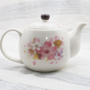 Pu&ograve; includere: Teiera in ceramica bianca con coperchio e manico. La teiera &egrave; decorata con disegni floreali rosa, oro e bianchi. Il coperchio ha un pomello marrone scuro. La teiera &egrave; su una superficie a righe bianche e argentate.