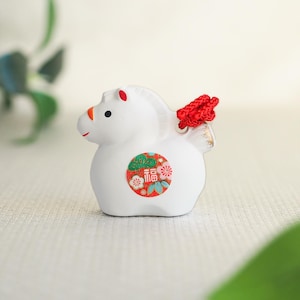 May include: A white ceramic horse figurine with red and orange accents. The figurine has a red braided detail on its back and a circular emblem with Japanese characters and floral designs.