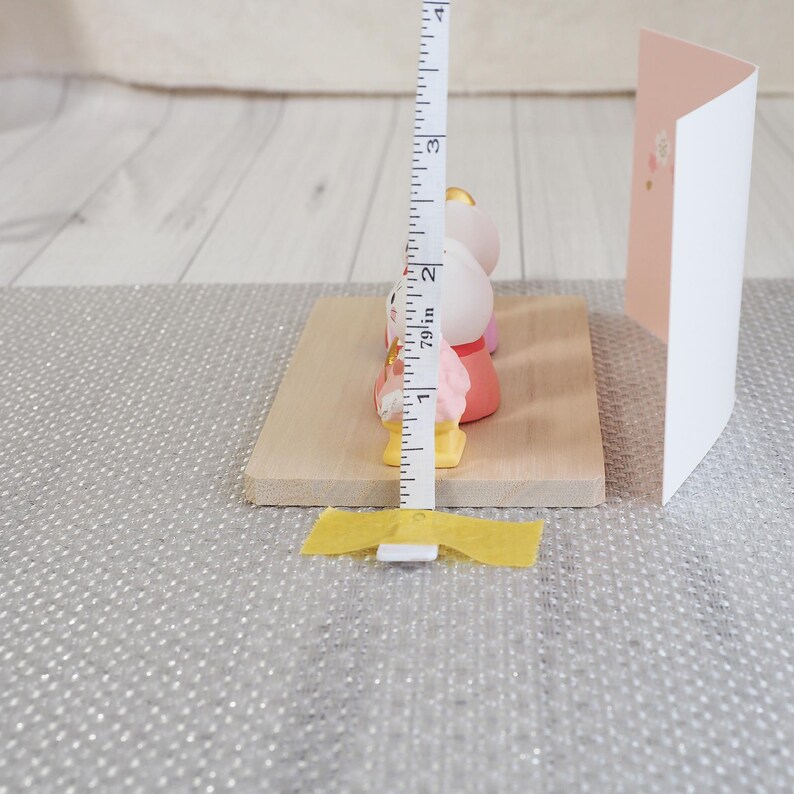 May include: A wooden base holds three small, colorful figurines, each approximately 2 inches tall. The figurines are pink, white, and yellow. A greeting card with a pink and white floral design stands to the right. A measuring tape is placed next to the figurines.