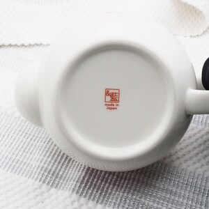 Pu&ograve; includere: Una teiera in ceramica bianca con un corpo arrotondato e un manico. Il fondo della teiera presenta un timbro rosso con testo giapponese che recita "made in Japan". La teiera &egrave; su una superficie strutturata.