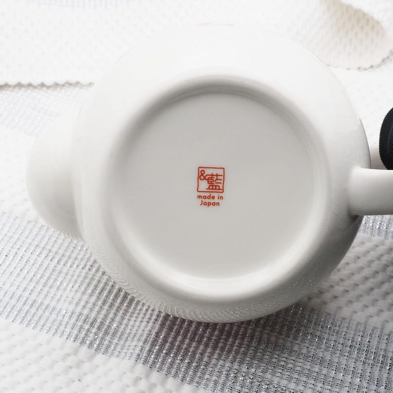 Pu&ograve; includere: Una teiera in ceramica bianca con un corpo arrotondato e un manico. Il fondo della teiera presenta un timbro rosso con testo giapponese che recita "made in Japan". La teiera &egrave; su una superficie strutturata.