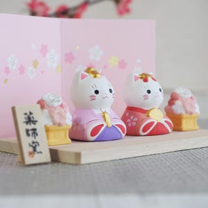 May include: A collection of Japanese cat figurines in traditional attire. The white cat figures wear colorful kimonos with gold accents. A small wooden sign and miniature desserts are also present, set against a pink backdrop.