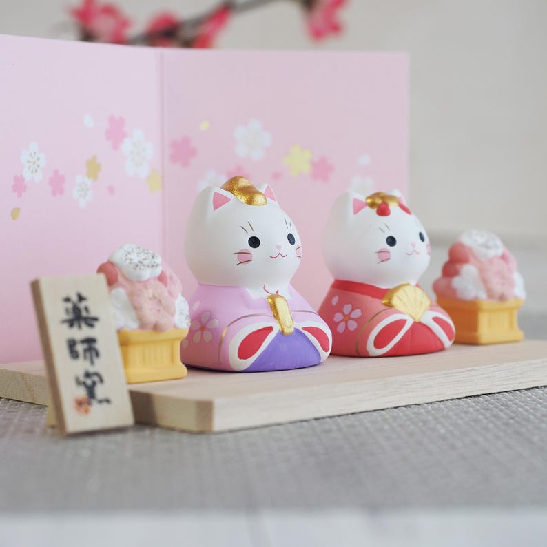 May include: A collection of Japanese cat figurines in traditional attire. The white cat figures wear colorful kimonos with gold accents. A small wooden sign and miniature desserts are also present, set against a pink backdrop.