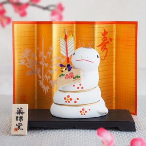 May include: A white ceramic snake figurine with red and gold accents, holding a sign with Japanese characters. It is set against a gold folding screen with a floral design and Japanese calligraphy. A small wooden sign is in front.