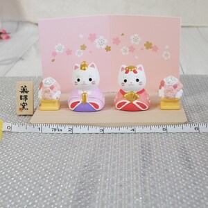 May include: A Japanese-style display featuring two cat figurines in traditional attire, flanked by decorative food items. The figurines are white with painted faces and wear pink, red, and purple garments. A wooden sign with Japanese text is on the left. The display is on a wooden base.
