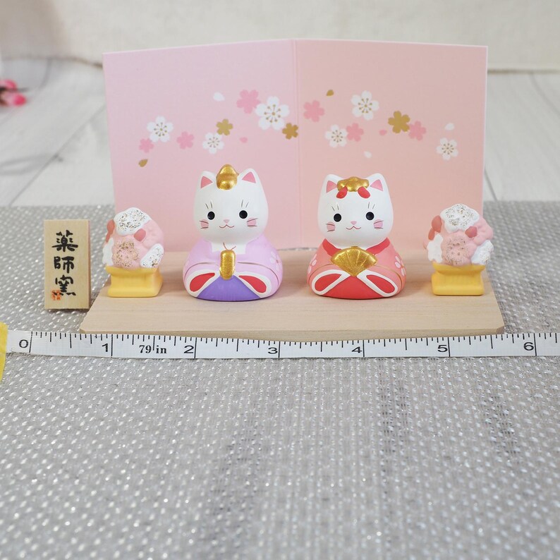May include: A Japanese-style display featuring two cat figurines in traditional attire, flanked by decorative food items. The figurines are white with painted faces and wear pink, red, and purple garments. A wooden sign with Japanese text is on the left. The display is on a wooden base.