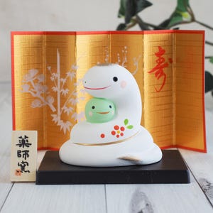 May include: A white ceramic snake figurine with a green frog nestled in its coils. The figurine has a smiling face and is decorated with red and gold accents. It sits on a black base in front of a gold screen.