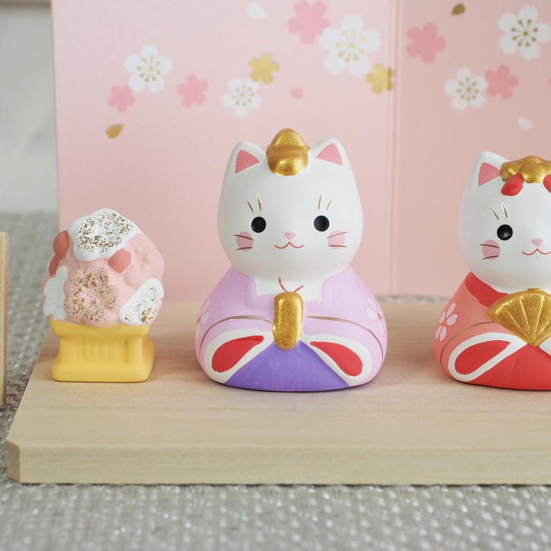 May include: A collection of Japanese ceramic cat figurines. The figurines are white with pink, purple, and gold accents. The background features a pink floral design. The figurines are displayed on a wooden surface.