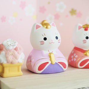 May include: Three ceramic cat figurines. The central cat wears a pink kimono with gold accents and a gold headpiece. The other cats are in similar outfits. The background is a soft pink with floral patterns. The figurines are on a wooden surface.