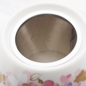 Pu&ograve; includere: Una teiera in ceramica bianca con un infusore a rete in acciaio inossidabile. La teiera presenta un design floreale in rosa, viola e oro. Il bordo superiore &egrave; bianco e l'infusore &egrave; visibile all'interno.