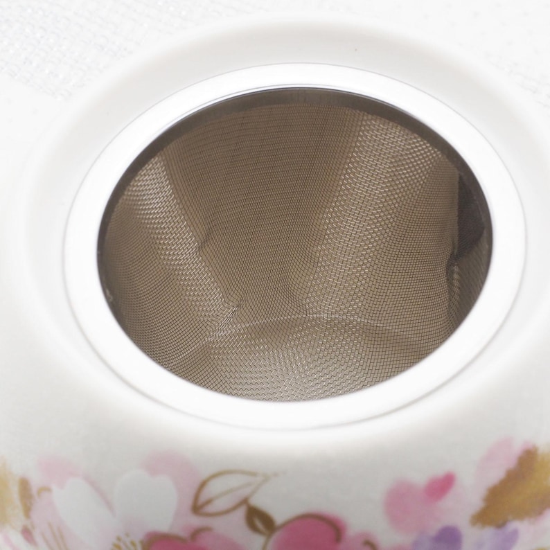 Pu&ograve; includere: Una teiera in ceramica bianca con un infusore a rete in acciaio inossidabile. La teiera presenta un design floreale in rosa, viola e oro. Il bordo superiore &egrave; bianco e l'infusore &egrave; visibile all'interno.
