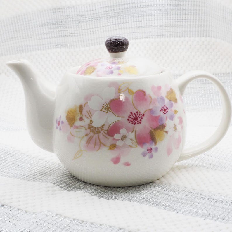 Pu&ograve; includere: Teiera in ceramica bianca con coperchio, decorata con motivi floreali rosa, oro e viola. La teiera ha un beccuccio e un manico curvi e un pomello marrone scuro sul coperchio. &Egrave; appoggiata su una superficie a righe bianche e argentate.