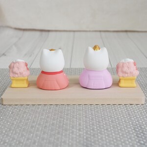 May include: Four small figurines on a wooden surface. Two cat-shaped figures wear pink and coral dresses with gold crowns. Two ice cream cone figures have pink ice cream and yellow cones. The background is a light gray surface.