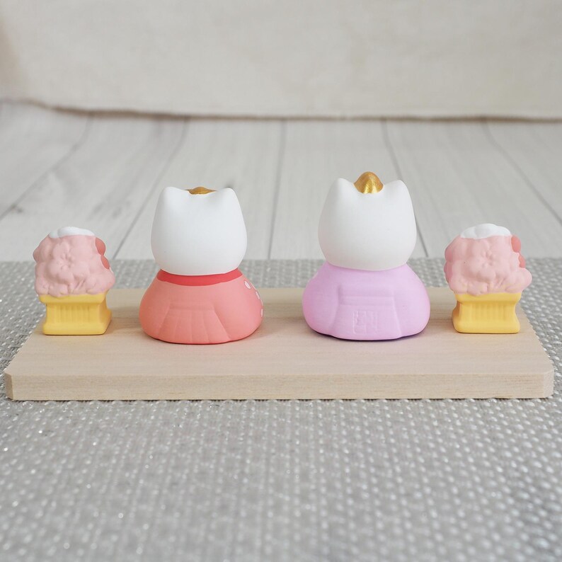 May include: Four small figurines on a wooden surface. Two cat-shaped figures wear pink and coral dresses with gold crowns. Two ice cream cone figures have pink ice cream and yellow cones. The background is a light gray surface.