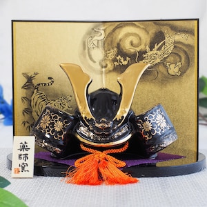 Pode incluir: Um capacete de samurai japonês decorativo com detalhes dourados e um borla laranja. O capacete preto apresenta detalhes dourados e está colocado sobre um fundo dourado com ilustrações de um tigre e um dragão. Uma pequena placa branca com texto japonês está na frente.