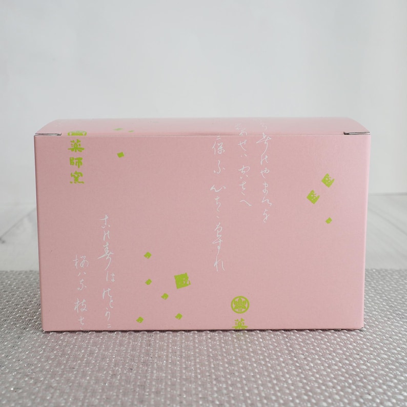 May include: A pink rectangular box with Japanese text and green geometric shapes. The box has text in Japanese on the side and top. The box is sitting on a textured, light gray surface.