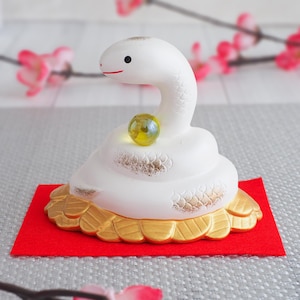 以下が含まれることがあります： 赤色のフェルトベースに金色の装飾が施された、白のセラミック製のヘビの置物。黄色と緑色のビー玉が特徴です。ヘビは赤い口と黒い目をしています。装飾品です。