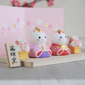 May include: A collection of Japanese cat figurines, dressed in traditional attire, with gold accents. The figurines are white with pink, red, and purple clothing. A small wooden sign with Japanese characters and a pink background with floral designs complete the scene.