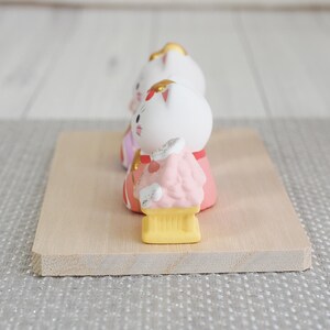 May include: Three ceramic cat figurines in traditional Japanese attire, arranged on a light wooden base. The cats have white bodies, pink and red clothing, and gold accents. The figurines are a charming representation of Japanese culture.