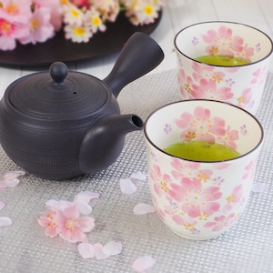 Könnte beinhalten: Eine dunkelgraue Teekanne mit seitlichem Griff und zwei weißen Teetassen, gefüllt mit grünem Tee. Die Teetassen sind mit rosa Kirschblütenmotiven verziert. Rosa Blütenblätter sind um die Tassen und die Teekanne verstreut.
