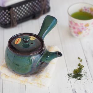 May include: A green ceramic teapot with a dark brown rim and handle, resting on a small, yellow-toned cloth. A cup of green tea and loose tea leaves are in the background. The teapot has a lid with a small knob and a sticker.