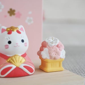 May include: A pair of Japanese-style figurines. One is a white cat with pink accents and a gold headpiece, wearing a red and gold outfit. The other is a small, pink and white dessert-shaped figurine on a yellow base.
