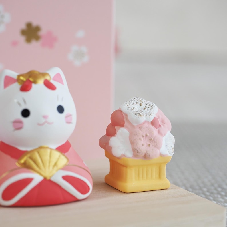May include: A pair of Japanese-style figurines. One is a white cat with pink accents and a gold headpiece, wearing a red and gold outfit. The other is a small, pink and white dessert-shaped figurine on a yellow base.