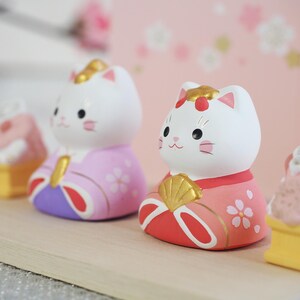 May include: Two white cat figurines with painted faces and traditional Japanese clothing. One cat wears a pink and purple kimono, the other a red and pink kimono. Both have gold accents and are decorated with floral patterns.