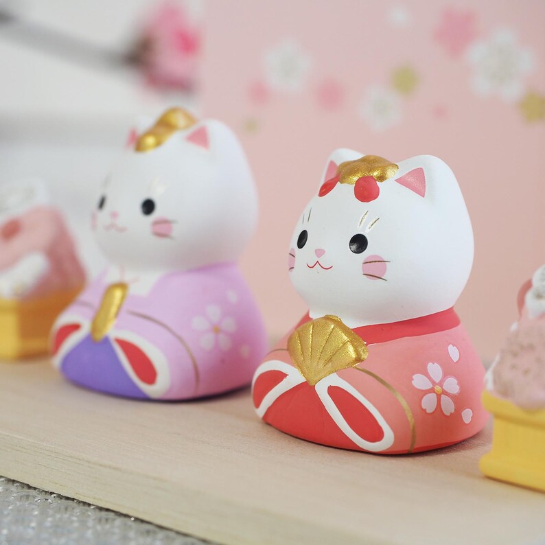 May include: Two white cat figurines with painted faces and traditional Japanese clothing. One cat wears a pink and purple kimono, the other a red and pink kimono. Both have gold accents and are decorated with floral patterns.