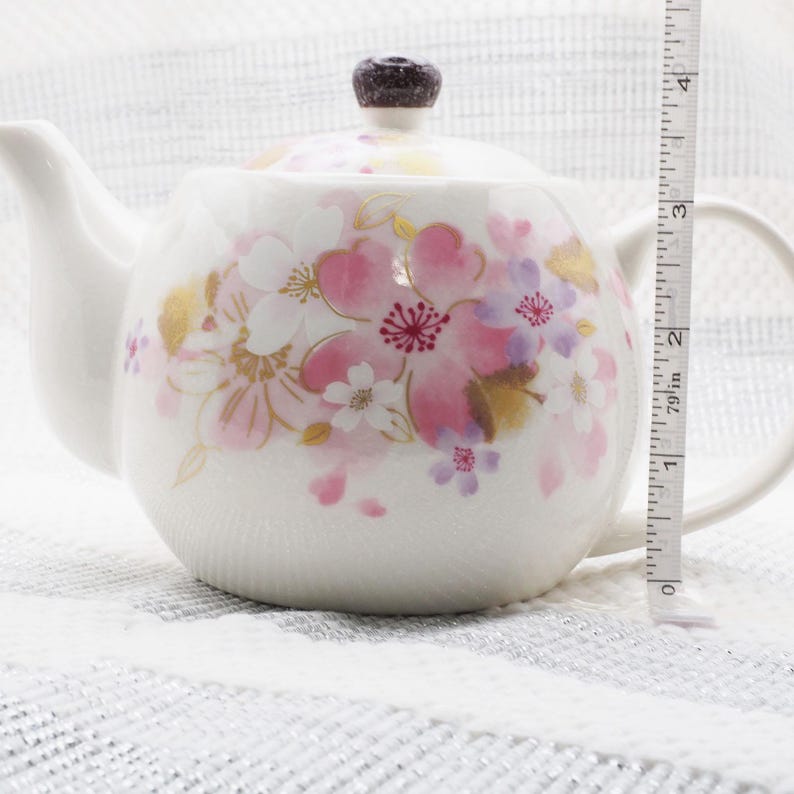 Pu&ograve; includere: Teiera in ceramica bianca con un design floreale con fiori rosa, bianchi e dorati. La teiera ha un pomello marrone scuro sul coperchio e un manico curvo. La teiera &egrave; alta circa 7.6 cm.