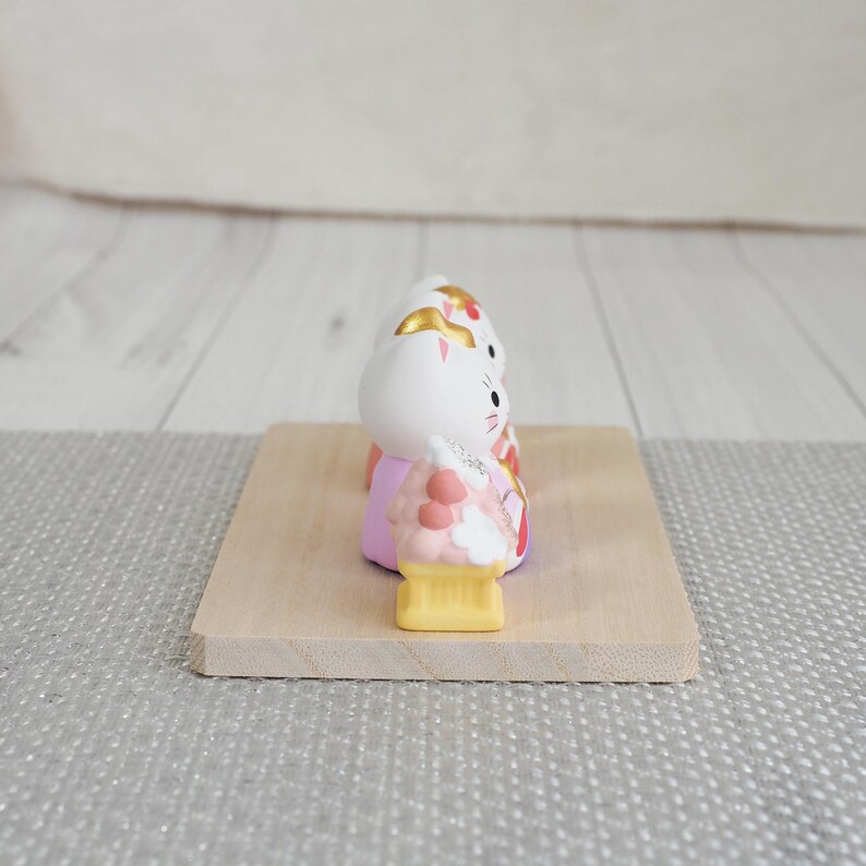 May include: A white ceramic Maneki Neko cat figurine with gold accents, a pink kimono, and a yellow base. The cat is on a small, light-colored wooden platform. The background is a light gray surface and a white wooden wall.