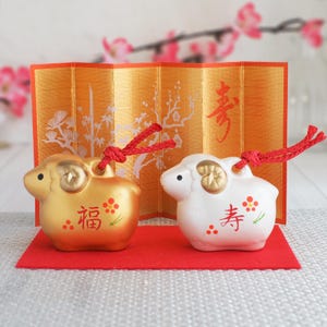 May include: Two ceramic sheep figurines, one gold and one white, each featuring a red cord loop, gold-painted horns, and red kanji characters with floral accents. They are displayed in front of a gold folding screen with a red base mat.