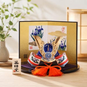 May include: A ceramic samurai helmet figurine featuring a glossy blue and white finish with gold accents, a central crest, and bright orange tassels. It is displayed on a black circular base in front of a gold folding screen with iris flower illustrations.