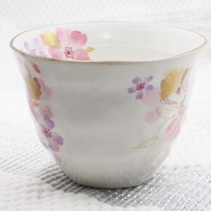Pu&ograve; includere: Una ciotola in ceramica bianca con una texture maculata, adornata da disegni floreali rosa, viola e oro. La ciotola ha una forma leggermente irregolare e un bordo dorato. Il motivo floreale comprende fiori di ciliegio.