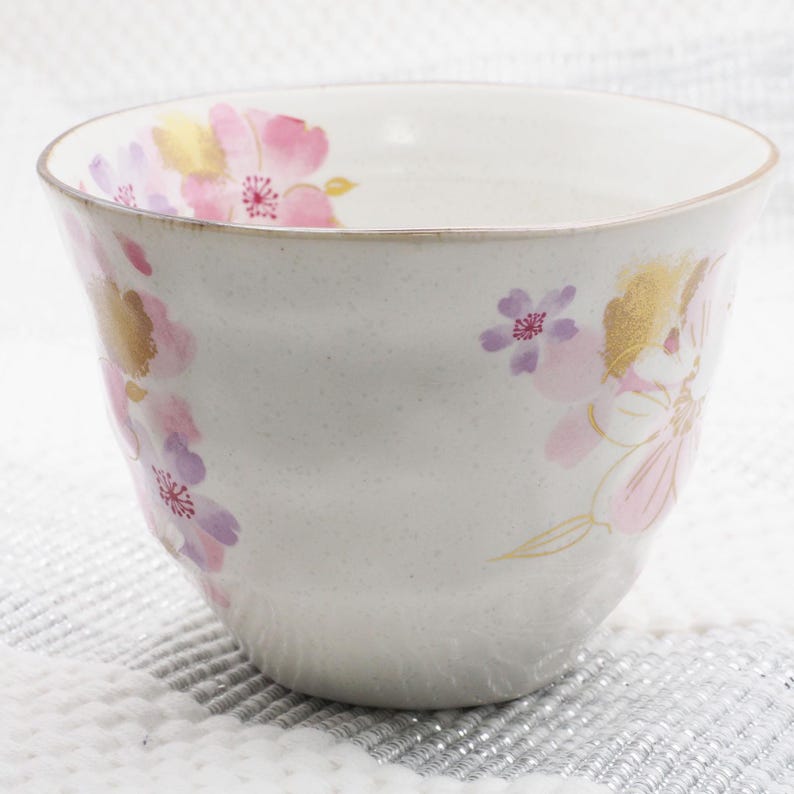 Pu&ograve; includere: Una ciotola in ceramica bianca con una texture maculata, adornata da disegni floreali rosa, viola e oro. La ciotola ha una forma leggermente irregolare e un bordo dorato. Il motivo floreale comprende fiori di ciliegio.