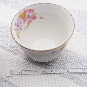 Pu&ograve; includere: Una tazza da t&egrave; in ceramica bianca con un motivo floreale rosa, oro e viola. La tazza ha una consistenza macchiettata e un bordo marrone. Un metro a nastro &egrave; posto vicino alla tazza.